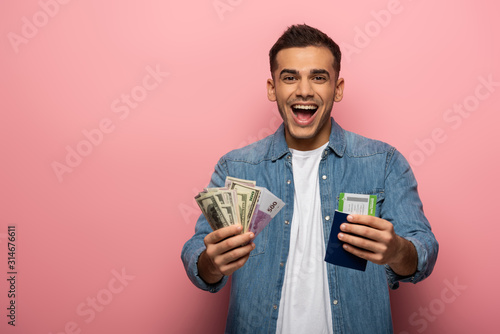 Surprised man holding currency, boarding pass and passport on pink background