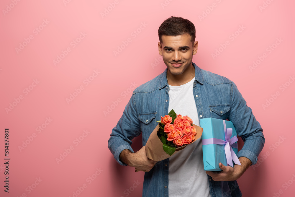 Handsome man holding gift and bouquet and looking at camera on pink background Handsome man holding gift and bouquet and looking at camera on pink background