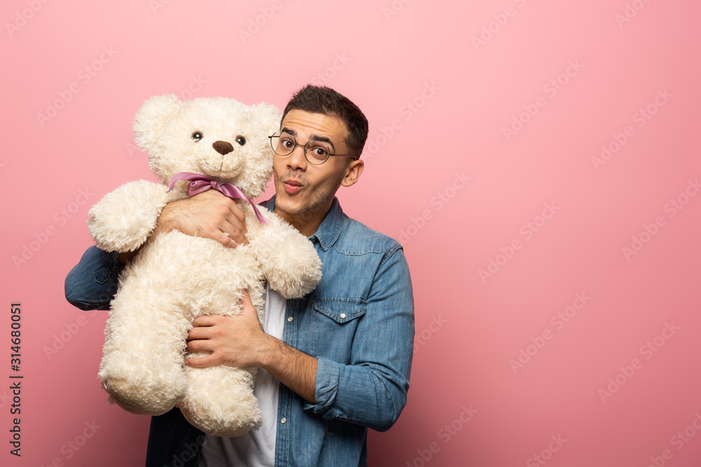 Young man grimacing at camera while holding teddy bear on pink background Young man grimacing at camera while holding teddy bear on pink background