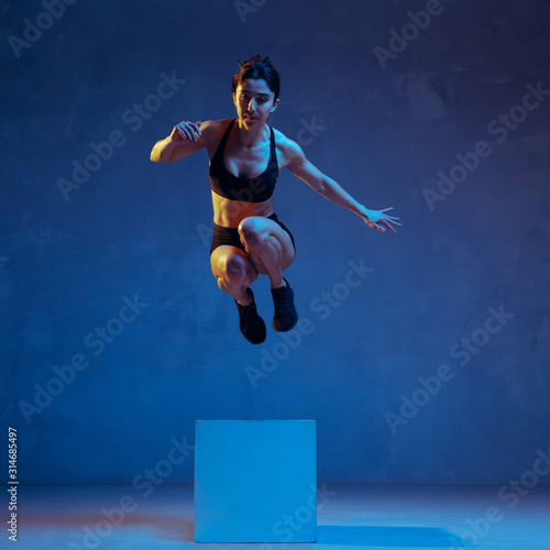 Fototapeta Naklejka Na Ścianę i Meble -  Caucasian young female athlete practicing on blue studio background in neon light. Close up of sportive model jumping on box. Body building, healthy lifestyle, beauty and action concept. Emotional.