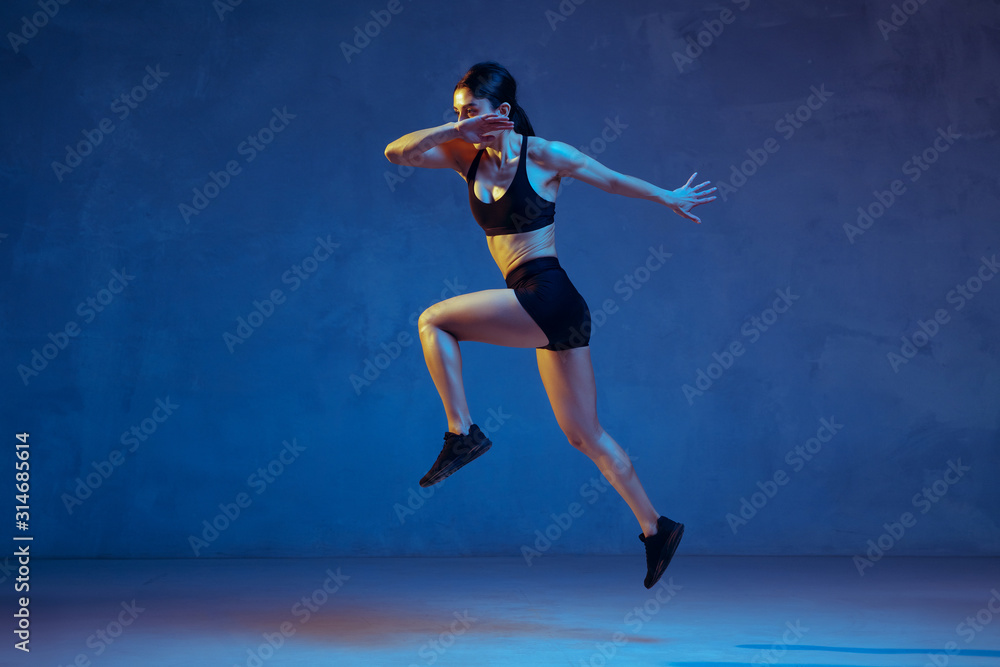 Caucasian young female athlete practicing on blue studio background in neon light. Close up of sportive model jumping high, running. Body building, healthy lifestyle, beauty and action concept.