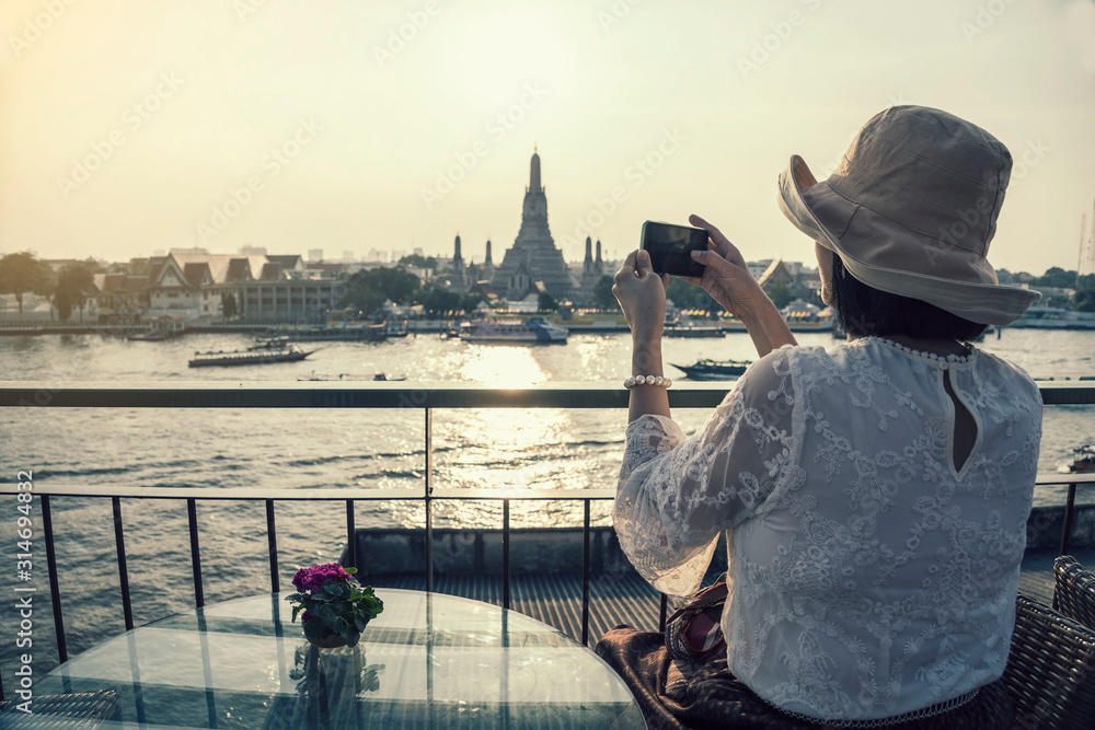 Asian tourists wearing hats, mobile phones, taking pictures Arun pagoda temple waterfront after sunset, The most famous tourist destination of Thailand