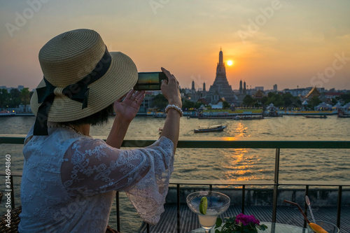 Asian tourists wearing hats, mobile phones, taking pictures Arun pagoda temple waterfront after sunset, The most famous tourist destination of Thailand