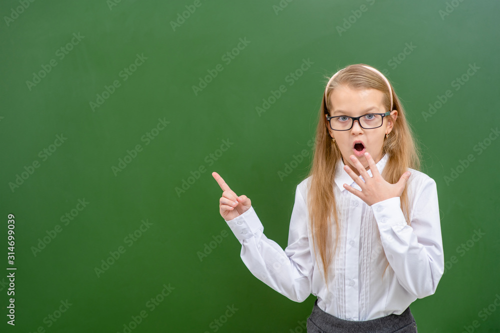 Shocked girl wearing eyeglasses points on empty chalk board. Empty space for text Shocked girl wearing eyeglasses points on empty chalk board. Empty space for text