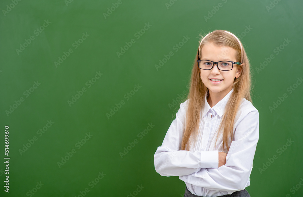 Young girl wearing a eyeglasses stands near green empty chalkboard. Empty space for text Young girl wearing a eyeglasses stands near green empty chalkboard. Empty space for text
