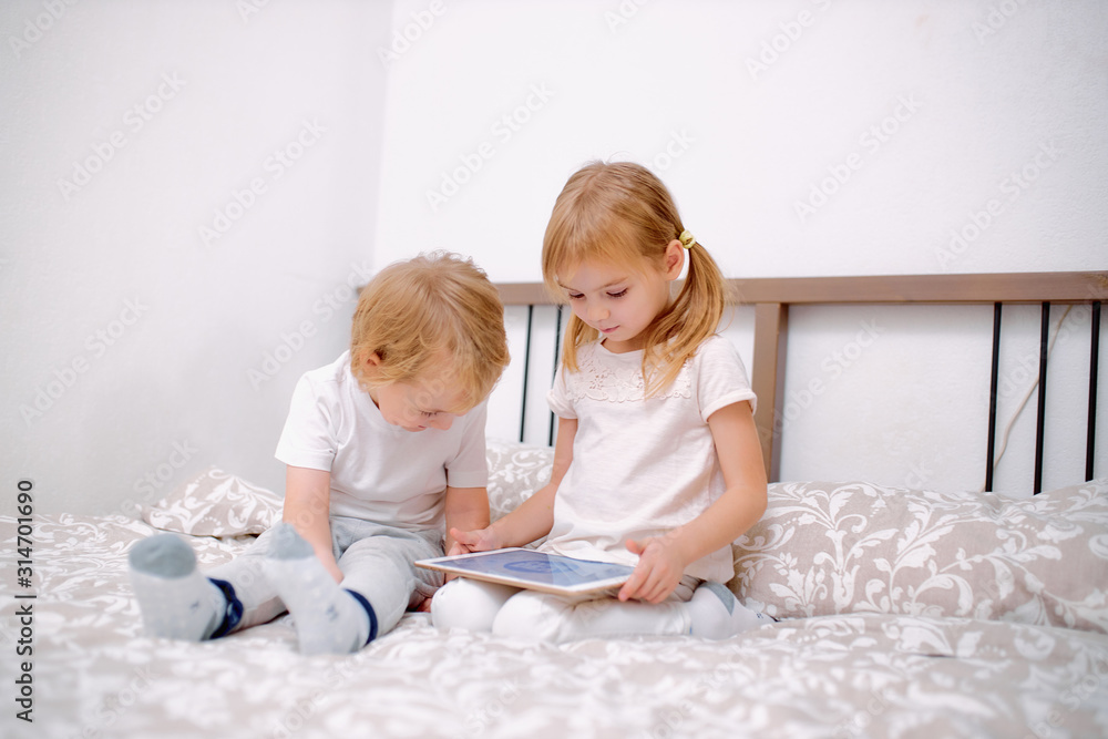 little caucasian children, boy and girl look at screen of laptop, playing or watching videos on modern gadget, wearing white casual clothes, sit on bed at home little caucasian children, boy and girl look at screen of laptop, playing or watching videos on modern gadget, wearing white casual clothes, sit on bed at home