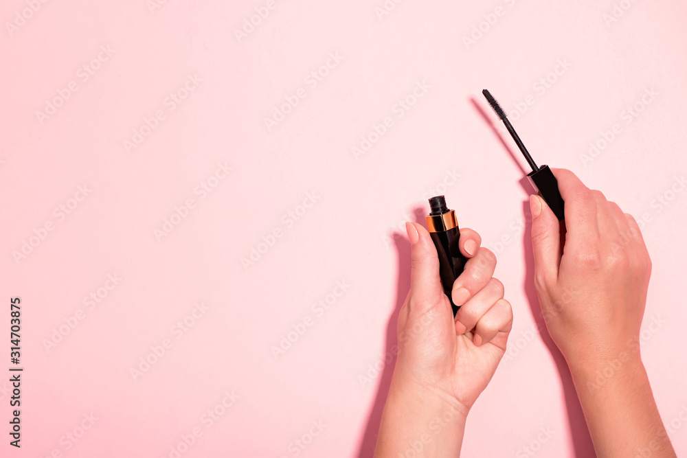 Woman hands holding open black mascara on pastel pink background, top view. Minimal beauty background with makeup products, flat lay, copy space. Eye make up concept Woman hands holding open black mascara on pastel pink background, top view. Minimal beauty background with makeup products, flat lay, copy space. Eye make up concept