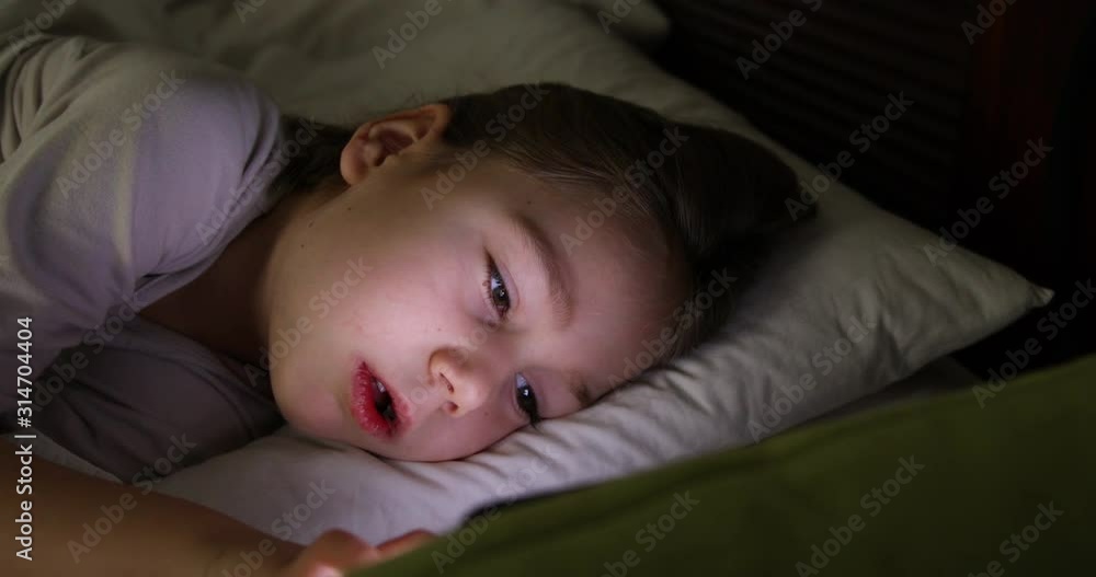 face lit of six years old girl typing on digital screen lying on bed at night face lit of six years old girl typing on digital screen lying on bed at night