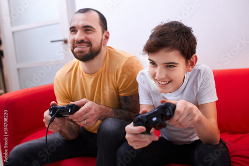 virtual, video game. caucasian bearded man playing video games with son indoors, sitting on sofa, wearing t-shirts, holding joysticks in hands. enjoy, leisure