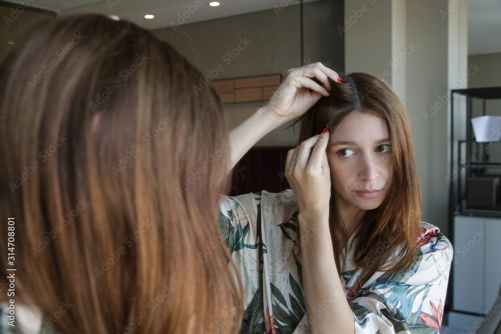 Naklejka premium Portrait of a beautiful young woman examining her scalp and hair in front of the mirror, hair roots, color, grey hair, hair loss or dry scalp problem