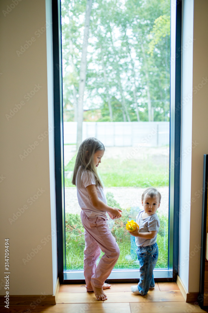 Children standing near panoramic window at home, baby surprisingly look at camera. Family concept Children standing near panoramic window at home, baby surprisingly look at camera. Family concept