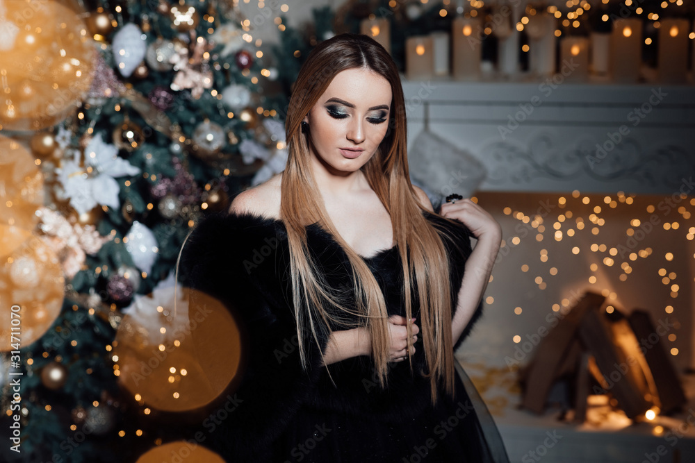 Pregnant woman hugging her belly. Happy young lady in elegant long black dress near the christmas tree. New year concept. Fashion interior holiday Christmas photo. Woman with evening make-up 