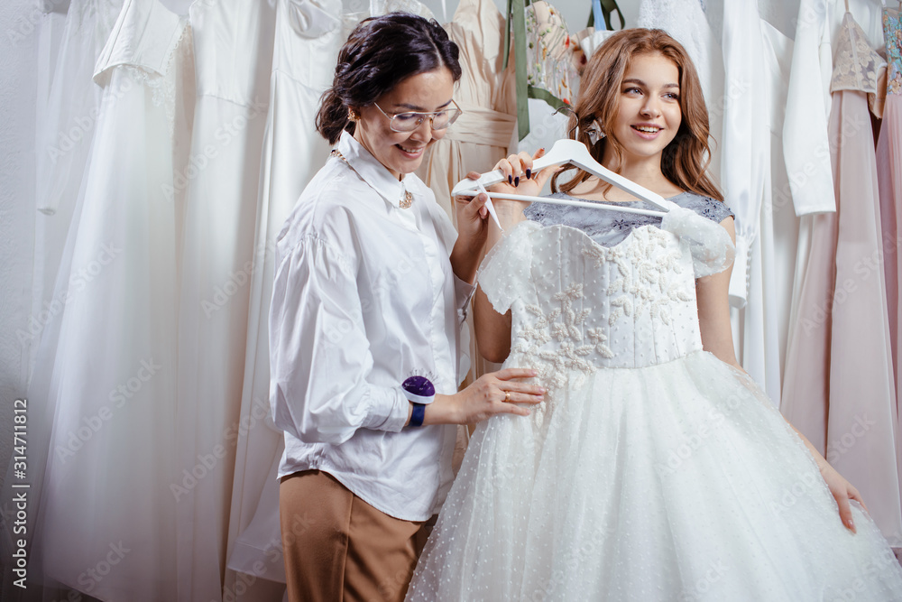 young beautiful caucasian bride going to buy wedding dress by designer in salon, try on it and look at mirror