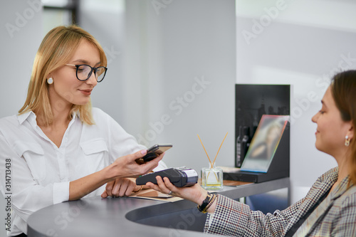 beautiful woman with blond short hair pay for beauty procedures in professional saloon, smiling friendly staff in reception photo