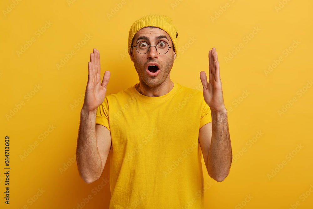 Stunned emotive man stretches hands and shows something huge, demonstrates large big size, tells about ammount, has shocked expression, dressed in casual wear, isolated over yellow background - obrazy, fototapety, plakaty 