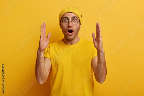 Stunned emotive man stretches hands and shows something huge, demonstrates large big size, tells about ammount, has shocked expression, dressed in casual wear, isolated over yellow background