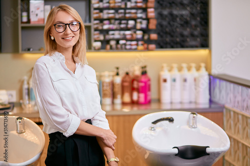 beautiful confident professional hairdresser in beauty saloon, caucasian woman with blond hair wearing white shirt stand looking at camera, isolated in beauty saloon