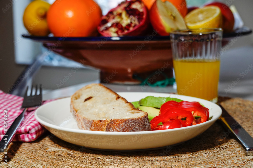 A piece of bread with avocado and peppers on a plate, with a ginger shot, and a fruit basket. A piece of bread with avocado and peppers on a plate, with a ginger shot, and a fruit basket.