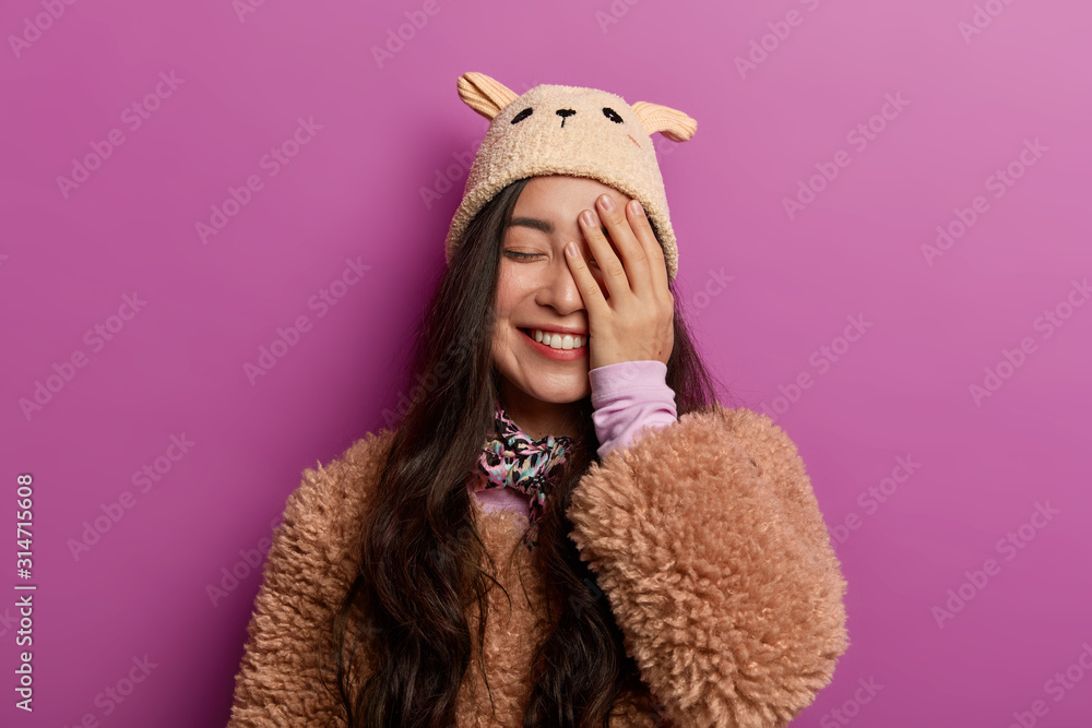 Positive sincere young woman laughs with joy, covers half of face, keeps eyes closed, dressed in winter outerwear, hears funny joke, isolated on lilac background. Emotions and feelings concept Positive sincere young woman laughs with joy, covers half of face, keeps eyes closed, dressed in winter outerwear, hears funny joke, isolated on lilac background. Emotions and feelings concept