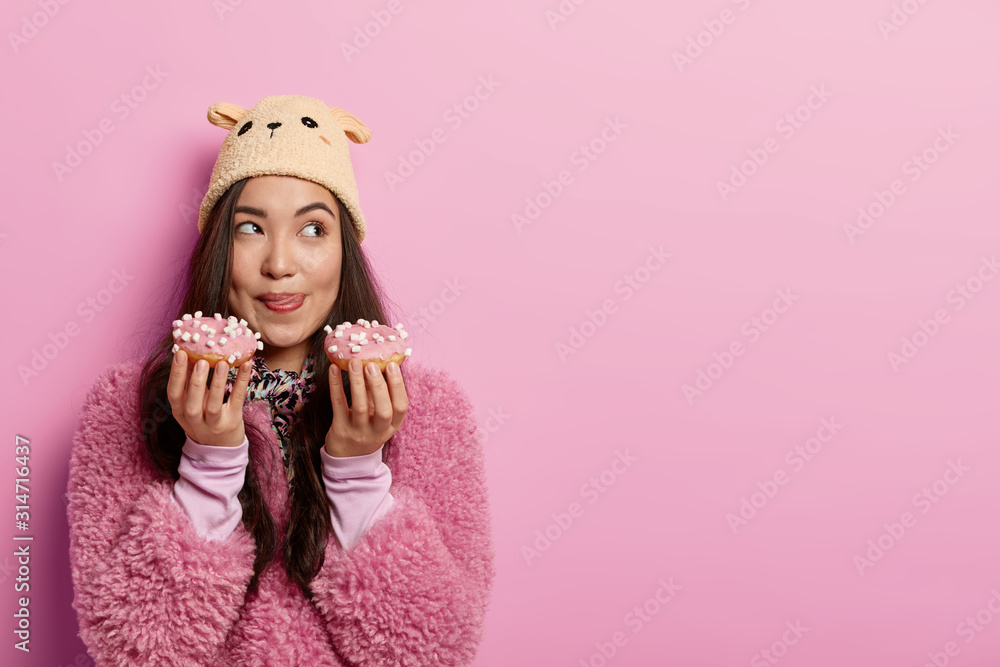 Dreamy young Asian lady holds two delicious sweet donuts, licks lips, wants to eat high calories food, breaks diet, dressed in warm clothes and headgear, stands indoor, blank space for your text Dreamy young Asian lady holds two delicious sweet donuts, licks lips, wants to eat high calories food, breaks diet, dressed in warm clothes and headgear, stands indoor, blank space for your text