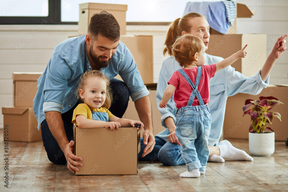 Caucasian family, man, woman and two girls play with moving box. Mother points to window of youngest daughter, father sat eldest daughter into box Caucasian family, man, woman and two girls play with moving box. Mother points to window of youngest daughter, father sat eldest daughter into box