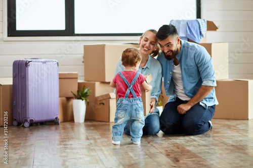 Kid wear denim overalls give little moving box parents who sit on floor and smile. Background moving boxes, suit and flower in pot