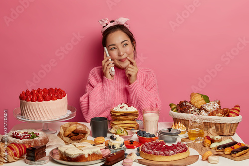 Yummy snacks. Pretty brunette Asian woman has phone conversation, invites friend to her home, feels temptation to eat tasty confectionery, wears knitted jumper, looks aside with happy expression