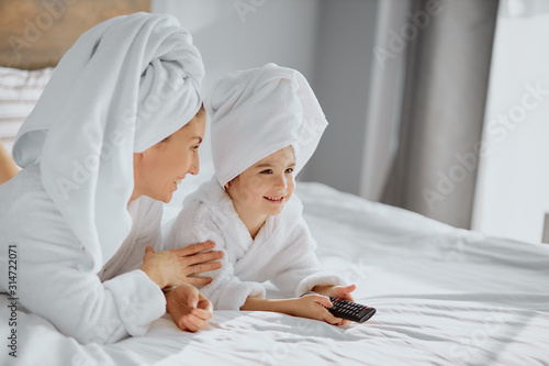 attractive woman and daughter sit holding tv remote going to watch tv on bed at home after shower