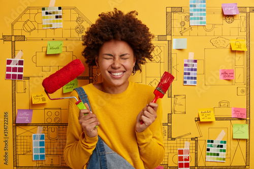 Photos Image of cheerful curly haired woman holds paint brush and roller, refurbishes w