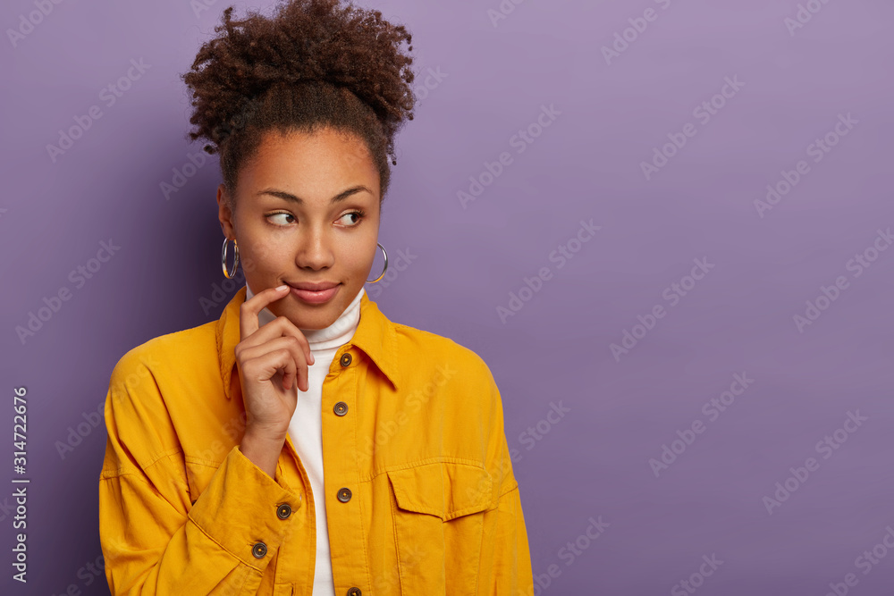Horizontal shot of thoughtful woman keeps finger near corner of lips, looks aside, thinks about making right decision, dressed in yellow clothing, isolated on purple wall. Pensive lady indoor