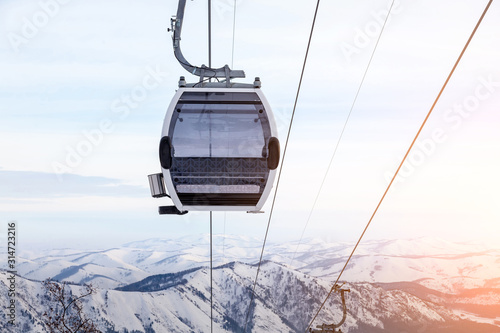 Cabin of a gondola cableway suspended on a rope where sits people with skis and snowboards high in the Altai mountains with snow and blue sky on winter sunset. Ski resorts and snowboarding.