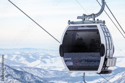 Cabin of a gondola cableway suspended on a rope where sits people with skis and snowboards high in the Altai mountains with snow and blue sky on winter sunset. Ski resorts and snowboarding. photo