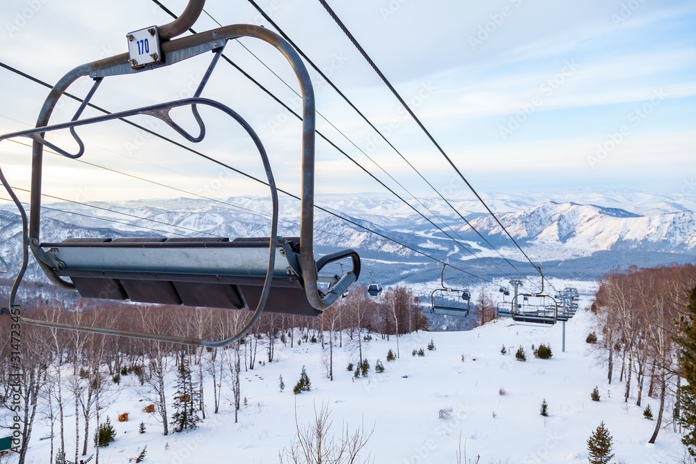 A ski lift chair cableway with a booth suspended on a cable in which sits people with skis and snowboards on a background of mountains, trees with snow on winter sunset. Ski resorts and snowboarding. A ski lift chair cableway with a booth suspended on a cable in which sits people with skis and snowboards on a background of mountains, trees with snow on winter sunset. Ski resorts and snowboarding.