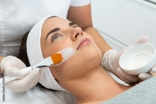 Kosmetolog wykonuje zabieg pielęgnacyjny cery. Nakłada krem na twarz kobiecie w salonie kosmetycznym. Kosmetyczka trzyma w dłoni pędzel i nakłada maskę na twarz. 