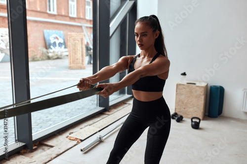 Young fit woman pull on herself elastic rope sports torso fitness biceps in gym. Dressed in black sportswear, window background