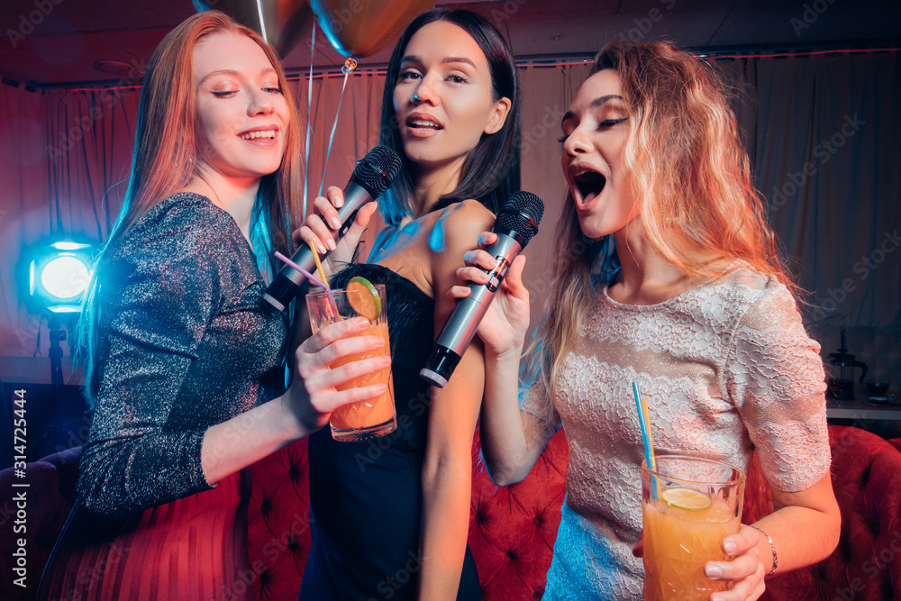 good-looking three female sing on microphone together in karaoke bar, wearing dresses and drinking juices, happy celebration