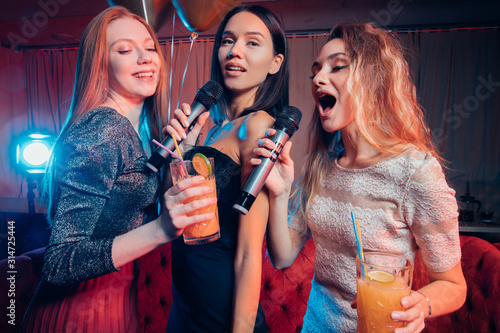 good-looking three female sing on microphone together in karaoke bar, wearing dresses and drinking juices, happy celebration