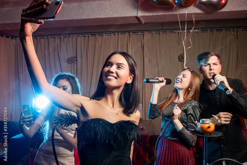 young and beautiful brunette lady in black dress take selfie with friends in karaoke bar, friends with balloons in the background, smoky space young and beautiful brunette lady in black dress take selfie with friends in karaoke bar, friends with balloons in the background, smoky space