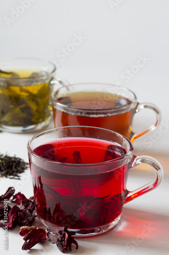 Red hot Hibiscus tea with dry flowers. Healthy lifestyle.Three glass cups with black, red and green tea on a white background. Green hot tea. Black hot tea. Hot drinks. Vertical orientation.