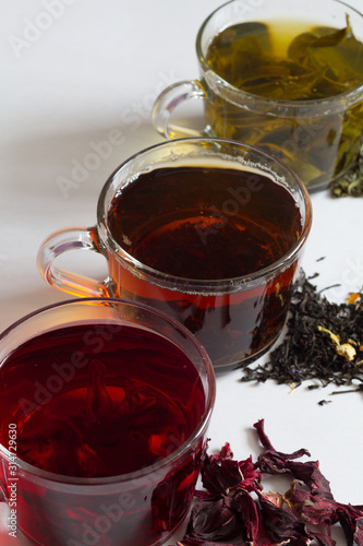 Three glass cups with black, red and green tea on a white background. Red hot Hibiscus tea with dry flowers. Healthy lifestyle.  Green hot tea. Black hot tea. Hot drinks.Vertical orientation.top view