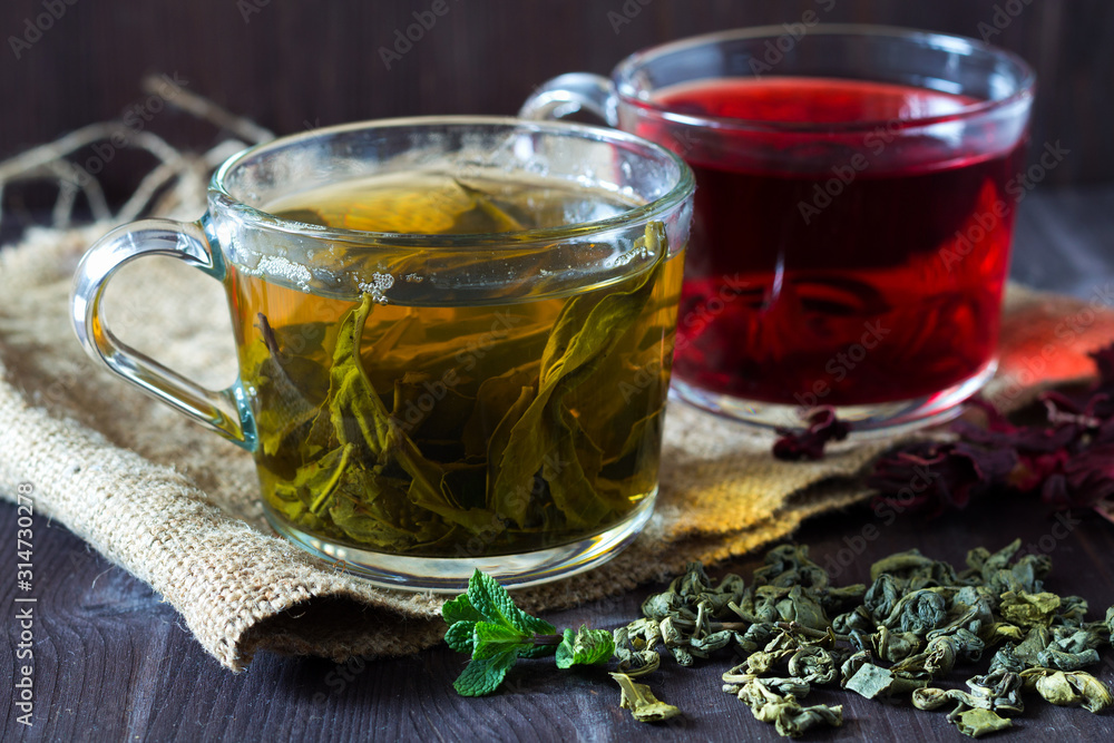 Healthy lifestyle. Glass cup of green herbal hot tea and cup of red hibiscus tea with dry leaves with fresh green leaves of mint with sackcloth on a dark wooden background. Hot drinks. Healthy lifestyle. Glass cup of green herbal hot tea and cup of red hibiscus tea with dry leaves with fresh green leaves of mint with sackcloth on a dark wooden background. Hot drinks.