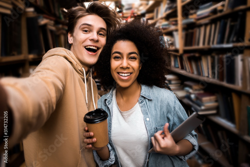 Positive happy selfie photo of two students friends multicultural couple in the library, happy faces and thirst for knowledge. Learning concept and student lifestyle.