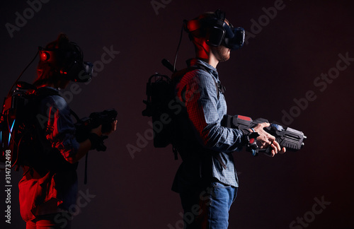 couple adventurers stand together back to back holding virtual guns, in fight. Isolated smoky background, red neon light