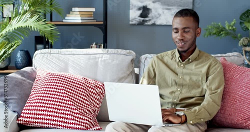 Happy smiled African American young guy typing on the keyboard of the laptop computerand smiling while sitting on the couch in the living room. photo