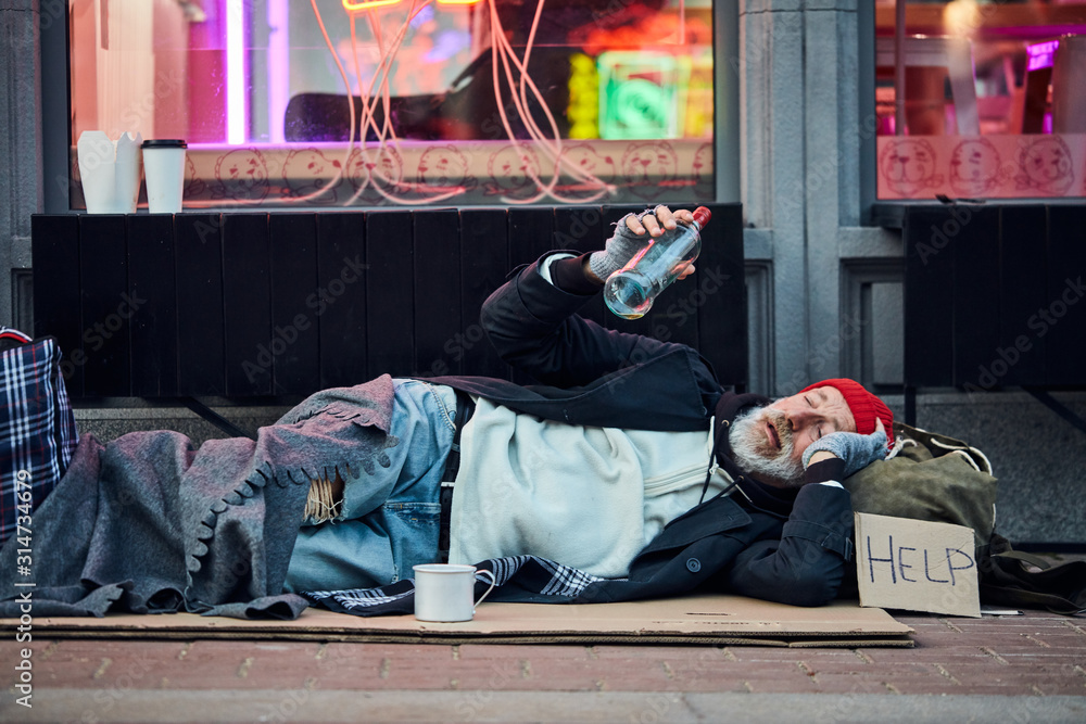 needy beggar male lying on cardboard box with sign HELP and bottle of alcohol, asking for help. Wearing old dirty clothes