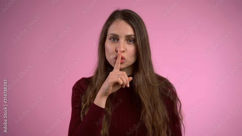 Beautiful long brown hair girl looking on camera and putting her finger on lips making silence gesture over pink background. Portrait of young attractive woman holding forefinger on lips and asking Beautiful long brown hair girl looking on camera and putting her finger on lips making silence gesture over pink background. Portrait of young attractive woman holding forefinger on lips and asking