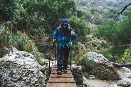 Man in blue jacket, with a backpack and walking sticks crossing a bridge in the mountain