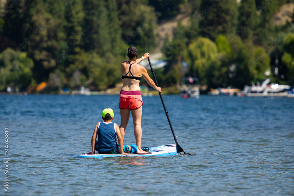 Obraz premium Back view of a young woman with son paddle boarding in sports clothing. Wide shot of mom and son during active holidays in sunny day