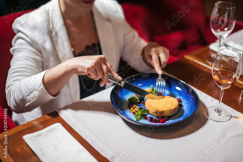 Tatented cook brought delicious dessert on glorious original plate, sweet cake prettified with various berris looks tasty on big blue plate. Woman in white jacket uses fork and knife to eat pastry