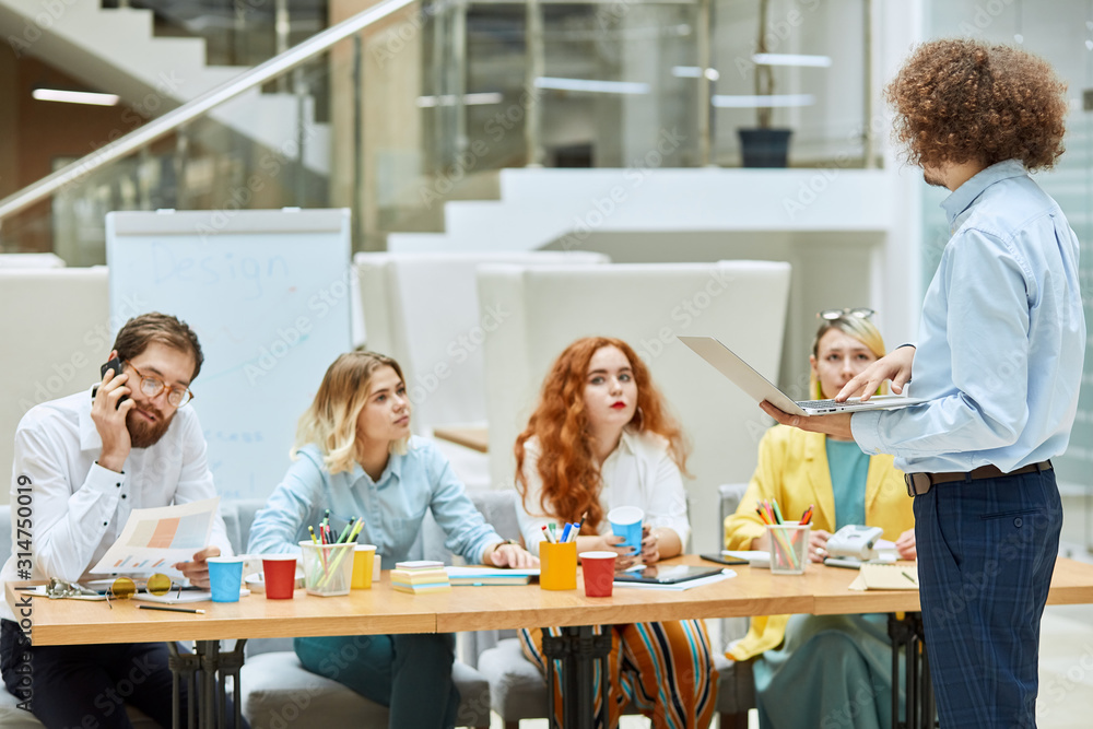 Tall, good shaped man with laptop computer in hands, has Afro hairstyle, turns towards young colleagues, tells important business issues in comfortable modern agency Tall, good shaped man with laptop computer in hands, has Afro hairstyle, turns towards young colleagues, tells important business issues in comfortable modern agency